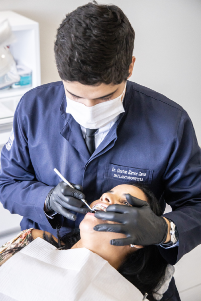 Doutor fazendo procedimento odontológico em uma cliente. Cliente deitada com papel de proteção sobre o busto e doutor manejando ferramenta odontológica com luvas pretas.