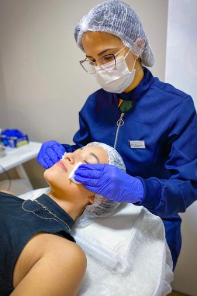 Doutora fazendo limpeza de pele com algodões em rosto de uma paciente. Paciente está deitada com olhos fechados e doutora utilizando luvas azuis e touca capilar para higiene.