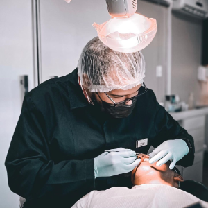 Procedimento dentário com um dentista manipulando boca de paciente. Dentista está com vestimenta preta, luvas azuis e com um equipamento de iluminação na direção da paciente.