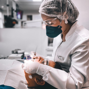 Procedimento dentário com uma dentista manipulando boca de paciente. Dentista está com vestimenta e luvas brancas. Foto tirada de lado, mostrando paciente deitada com dentista de perfil.