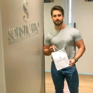 Cliente satisfeito posando ao lado da Logo da Sofisticatto na Clínica de Estética. Cliente está segurando presente de mimo da Sofisticatto.